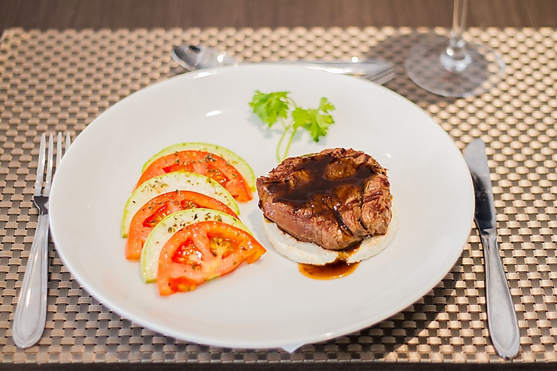 A plate with steak, tomato slices, and a garnish on a table with a woven placemat.
