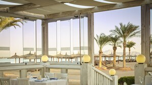 Un área de comedor al aire libre con mesas y sillas, con vista a una playa con palmeras y sombrillas de paja.
