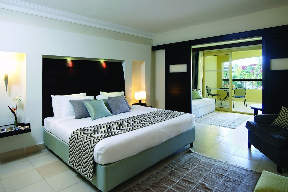 Quarto de hotel moderno com uma cama grande, área de estar com sofá, varanda com cadeiras e vista para o verde.