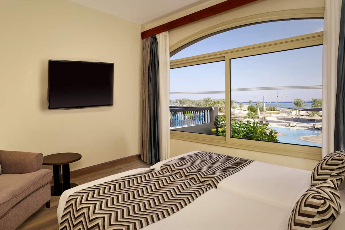 A hotel room with a large bed, a sofa, a small table, a TV, and a window with a view of a resort and pool.