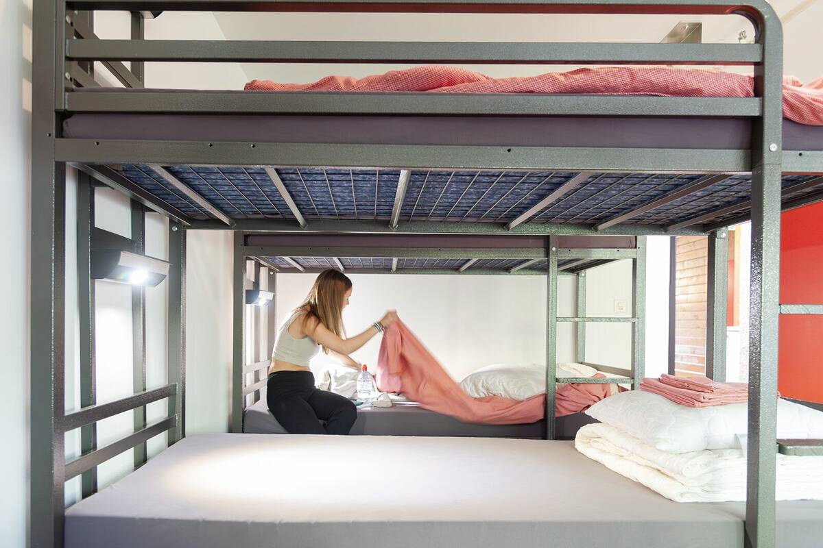 Una cama litera con una persona acomodando una manta, una ventana y una puerta.