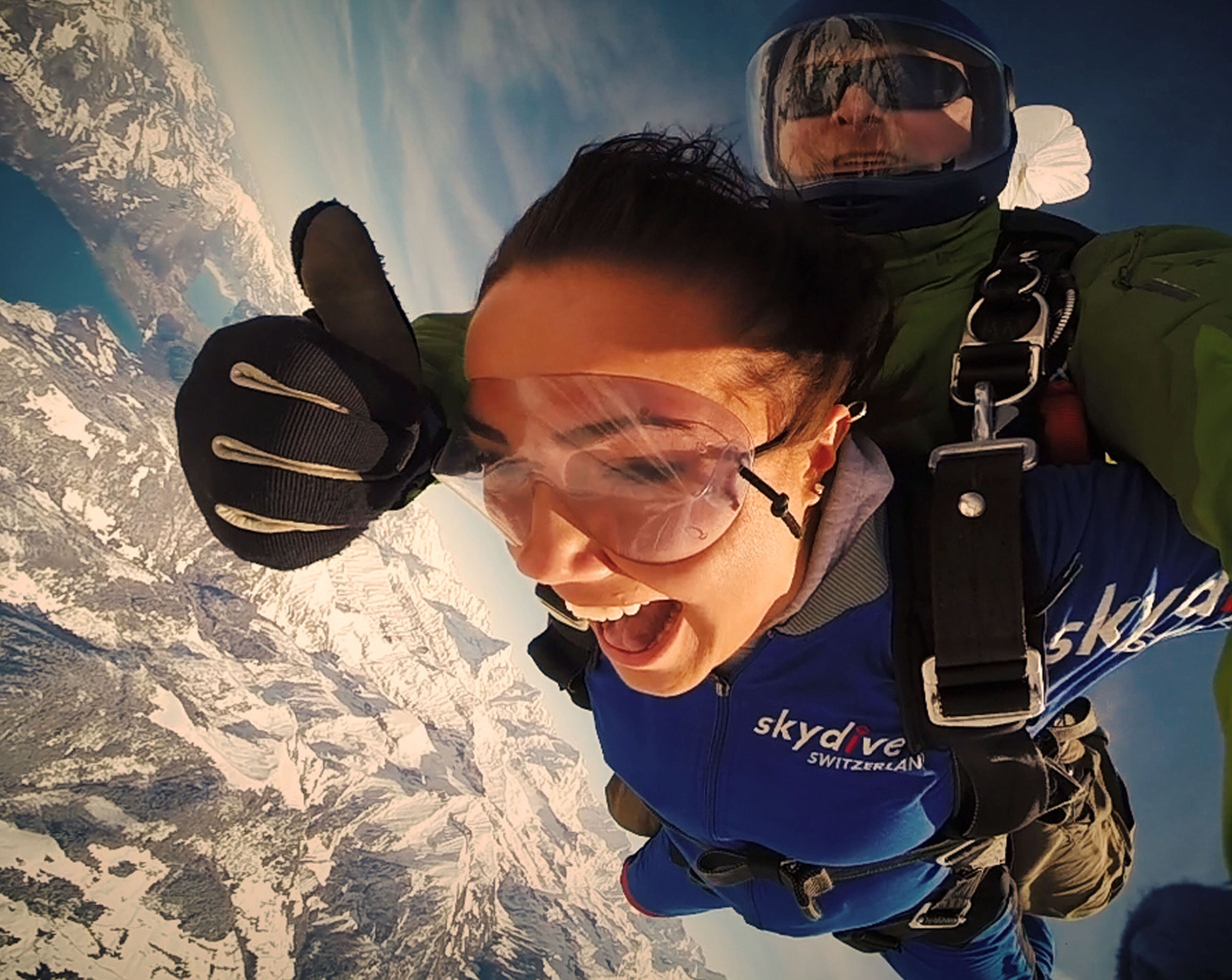 Una persona haciendo paracaidismo con un paracaídas, vestida con un traje de paracaidismo azul que lleva la frase 'skydiving Switzerland'.