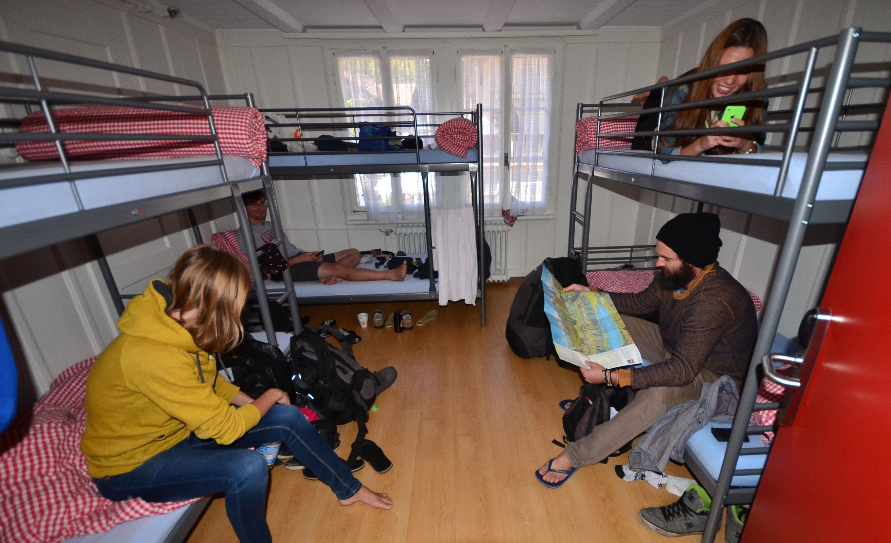 A dormitory room with bunk beds, a wooden floor, and a window with curtains.