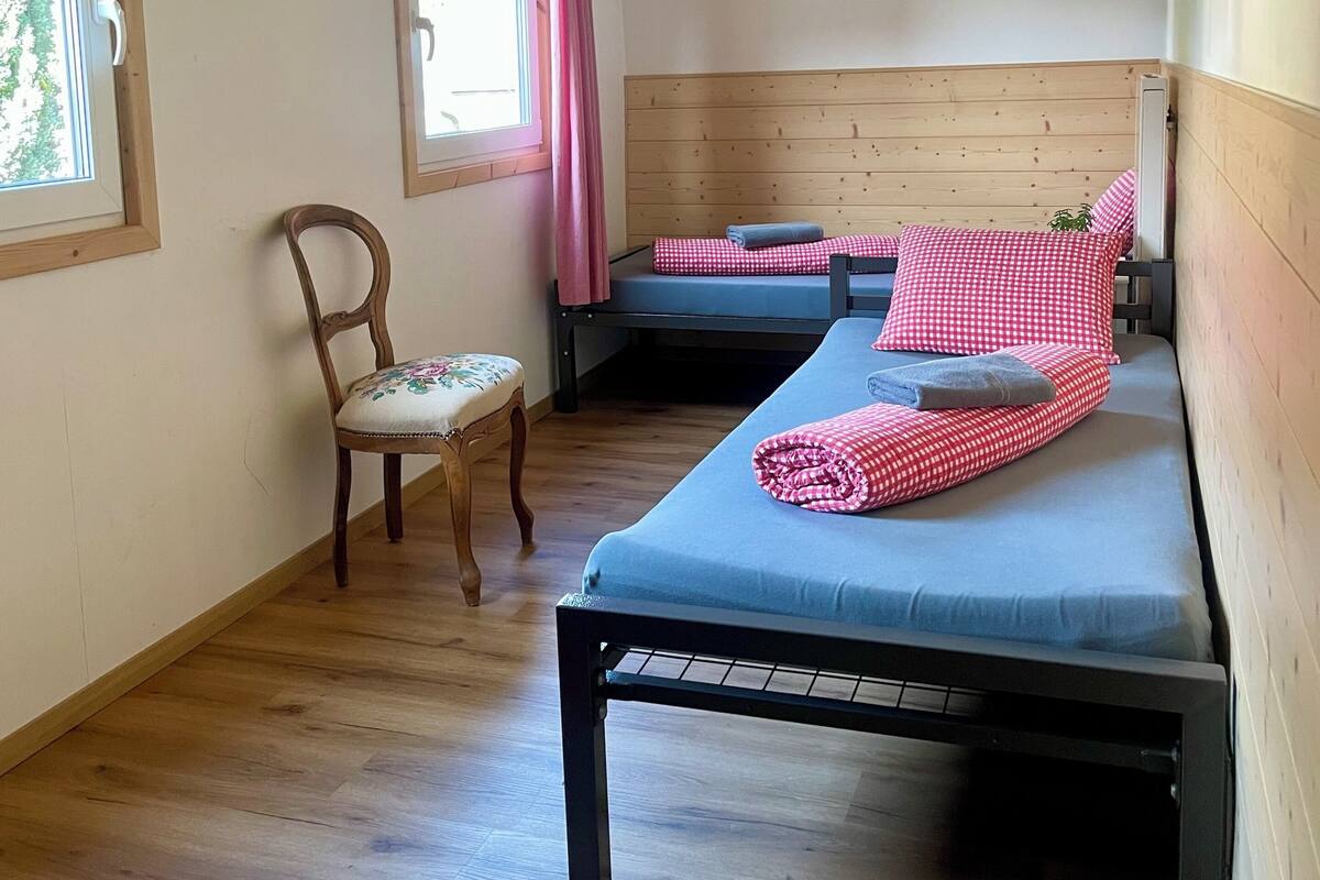 Una habitación con techo de madera, dos camas con ropa de cama a cuadros, una silla de madera, una ventana con cortina y una alfombra roja en el suelo.
