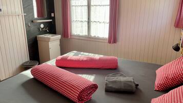 A bedroom with a bed, pillows, a small bedside table, a mirror, a window with curtains, and a wall-mounted shelf.
