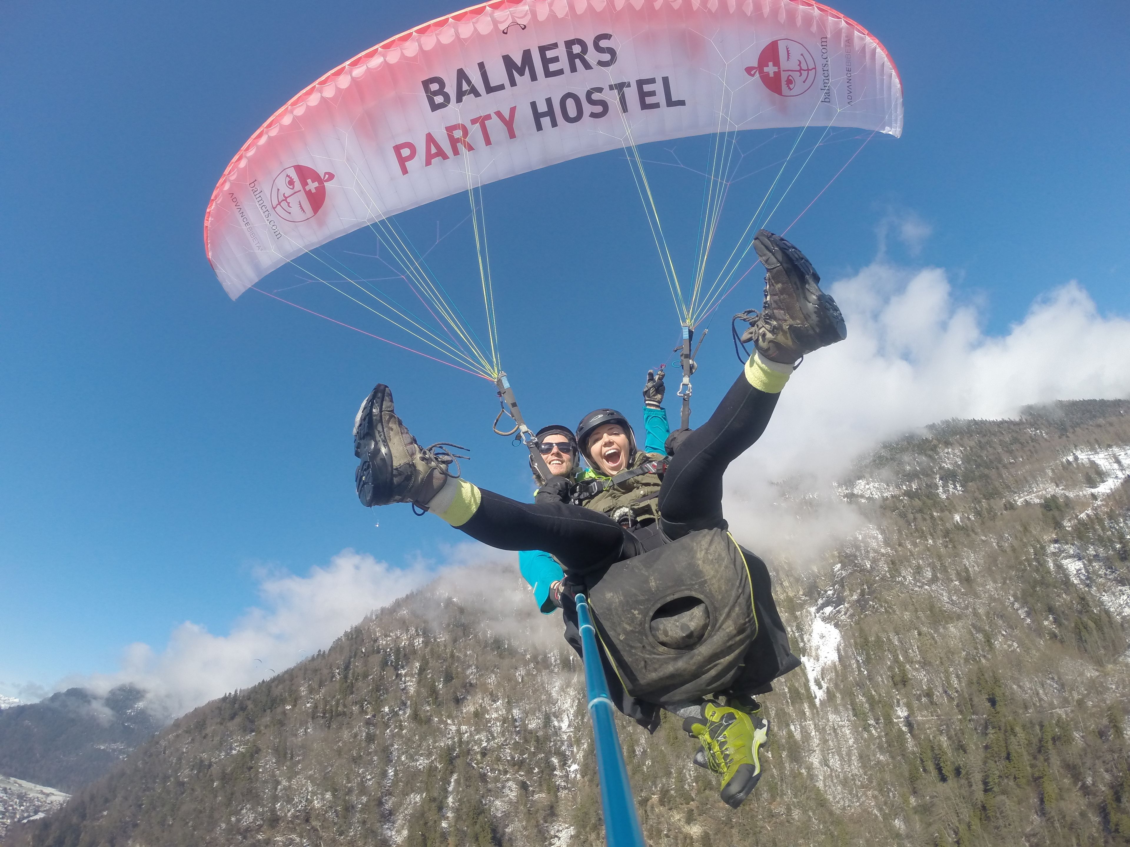 Une personne pratiquant le parapente avec une banderole sur laquelle on peut lire « BALMERS PARTY HOSTEL », avec un paysage montagneux en arrière-plan.