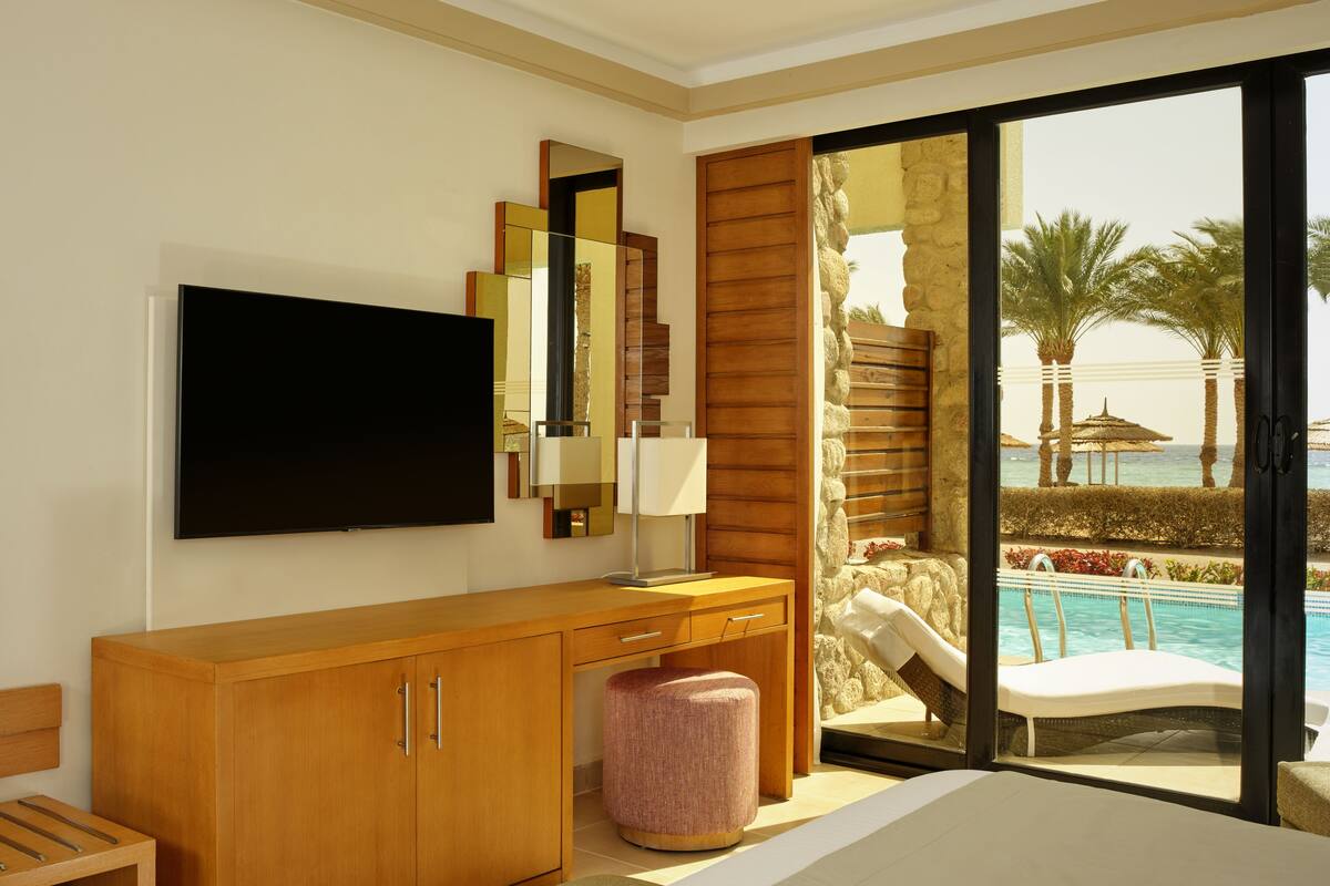 A hotel room with a TV, wooden furniture, a small pink ottoman, a pool view, and a stone wall.