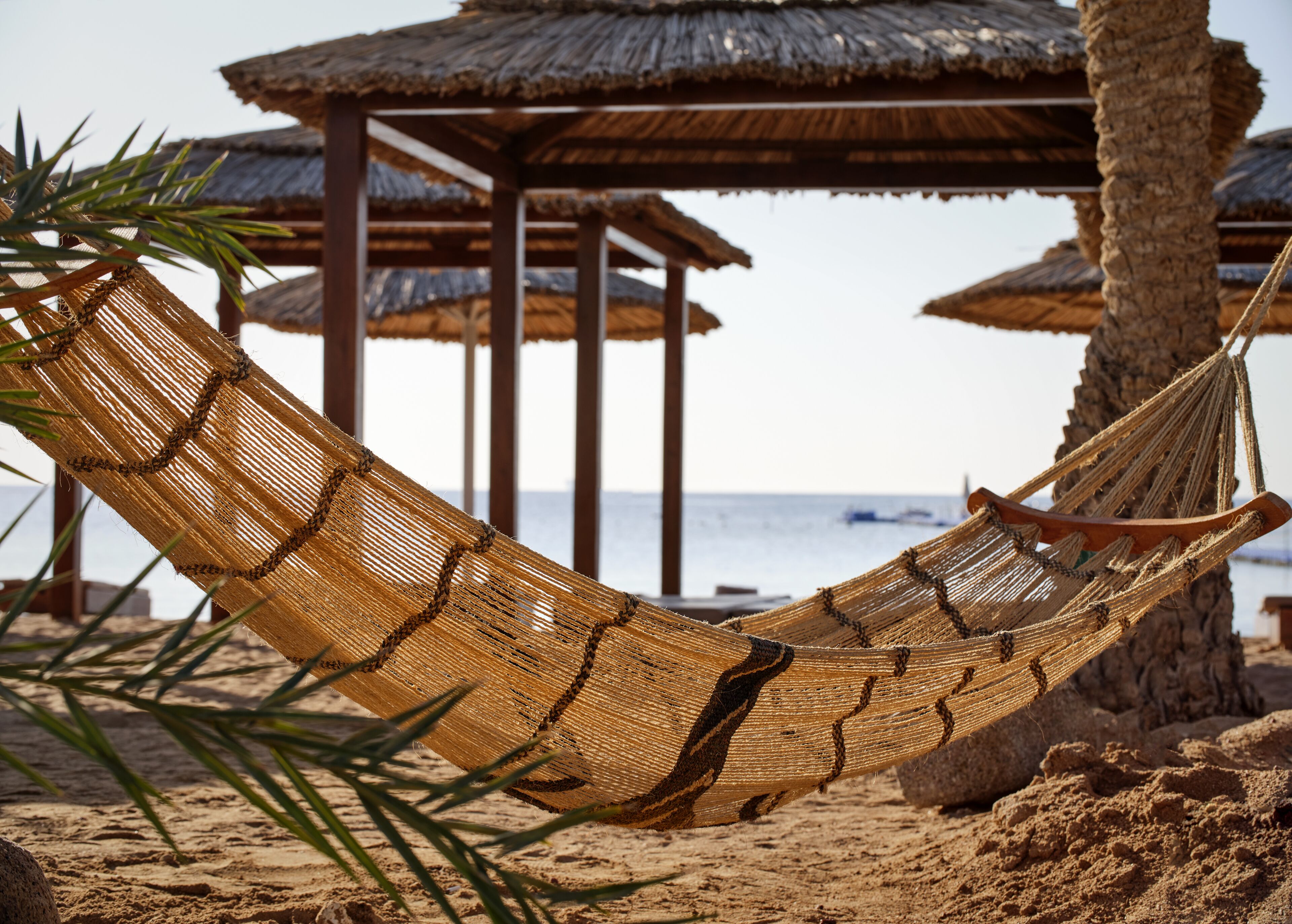 Una hamaca en una playa arenosa con sombrillas de techo de paja al fondo.