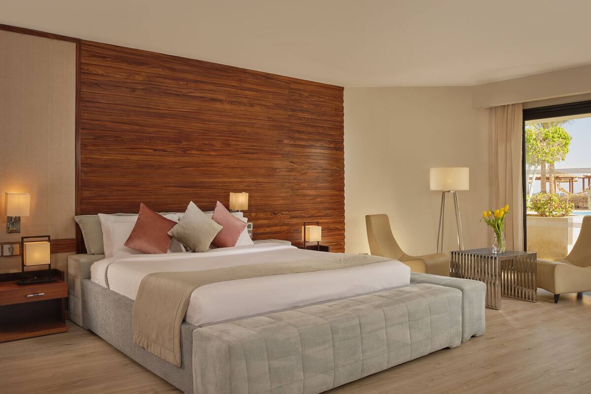 A modern hotel room with a large bed, wooden headboard, two bedside tables with lamps, a small side table with a vase, and a view of the pool area.