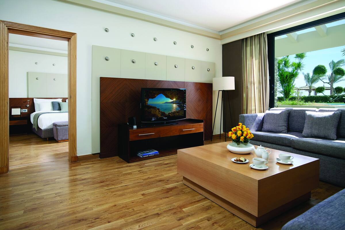 A hotel room with a wooden floor, a television on a wooden cabinet, a sofa, a coffee table with cups and a flower vase, and a view of palm trees through the window.