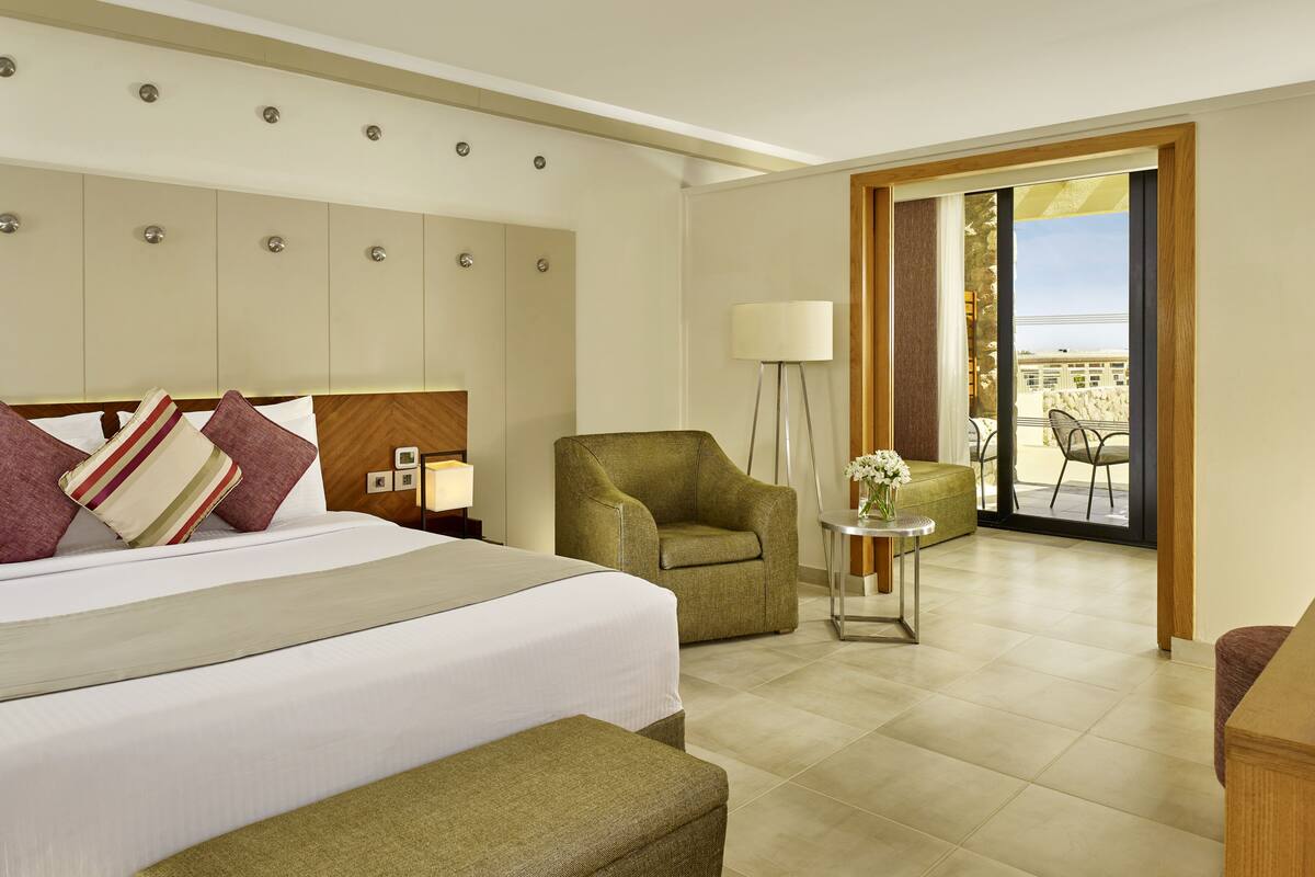 A hotel room with a bed, a green armchair, a small table with a lamp, and a balcony with outdoor furniture.