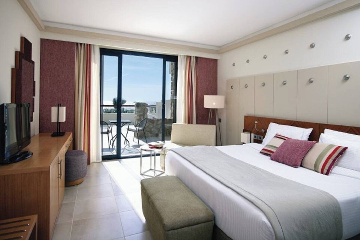 A hotel room with a bed, a desk with a TV, a chair, a lamp, and a view of the outdoors.