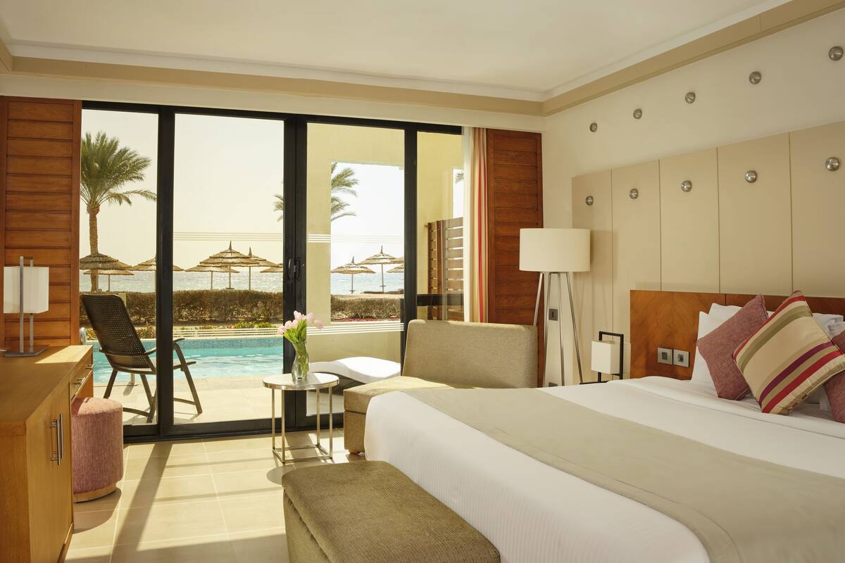 A hotel room with a bed, a desk, a chair, a small table, and a view of a pool and palm trees.