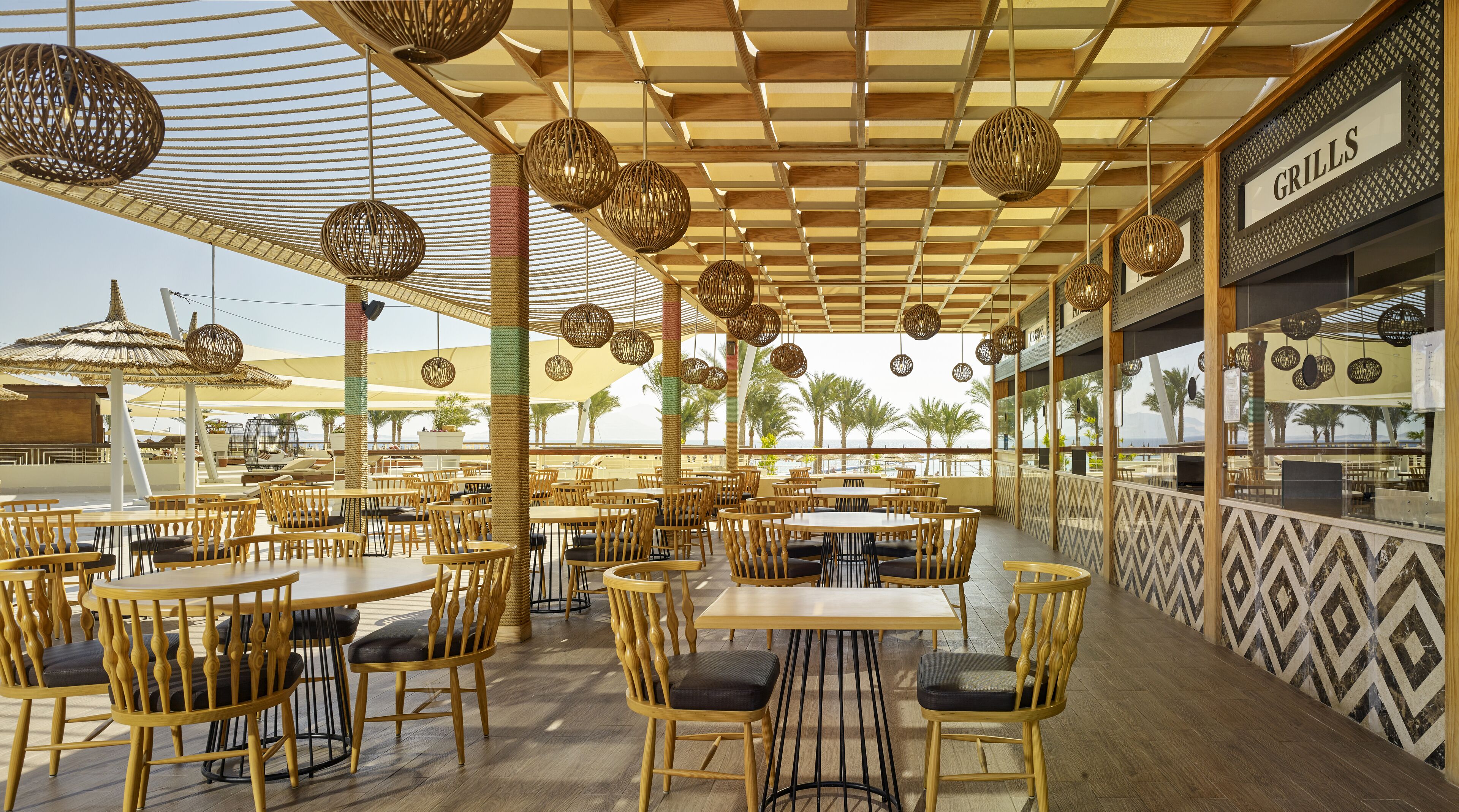 An outdoor dining area with wooden tables and chairs, hanging wicker lamps, and a view of the beach.