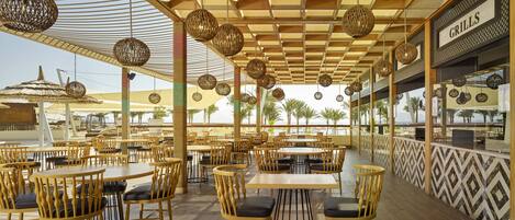 An outdoor dining area with wooden tables and chairs, hanging wicker lamps, and a view of the beach.