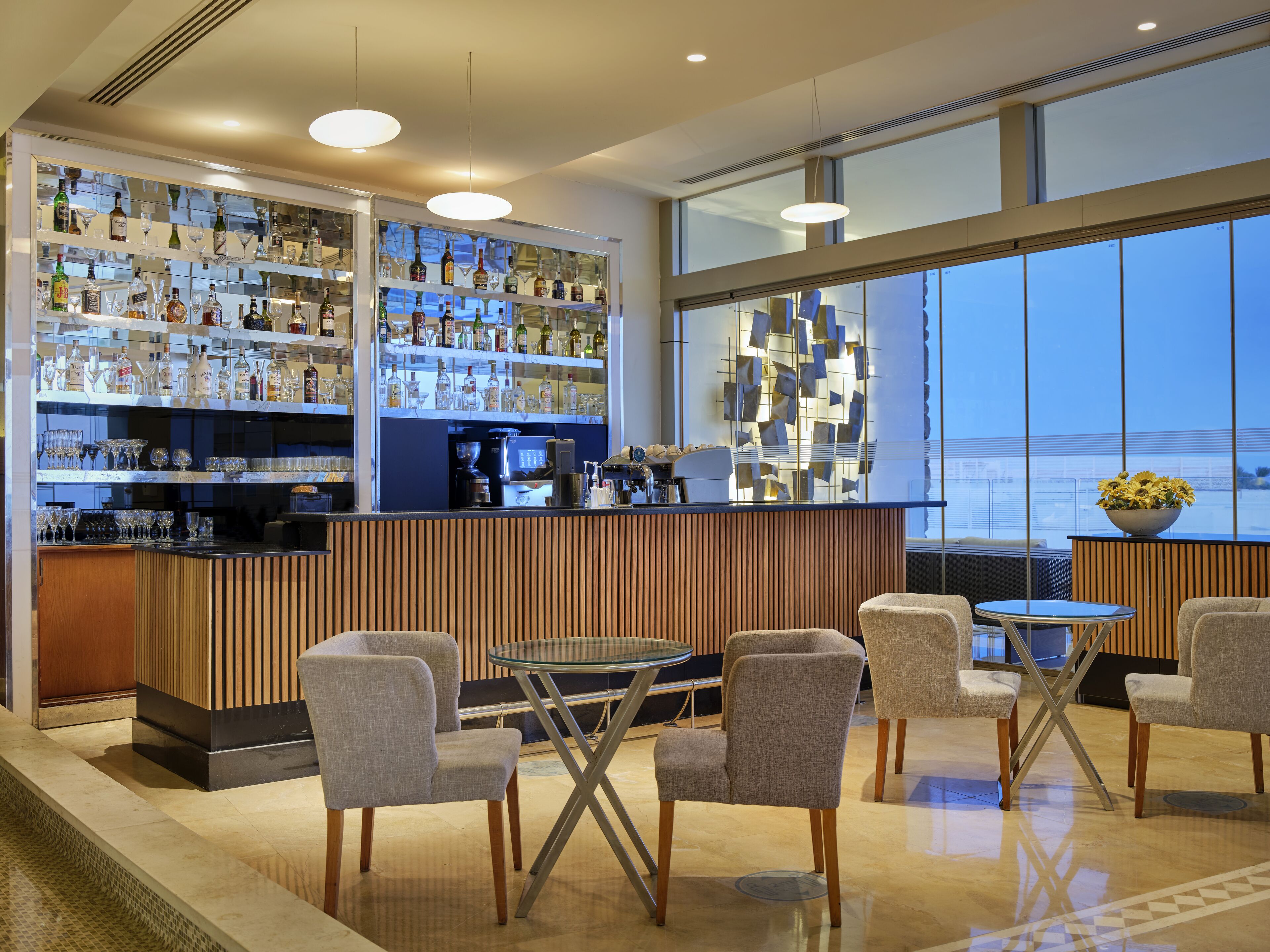 Un bar moderno con mostrador de madera, varias estanterías llenas de botellas de licor y vasos, y una máquina de café. Hay cuatro sillas tapizadas y una pequeña mesa redonda.