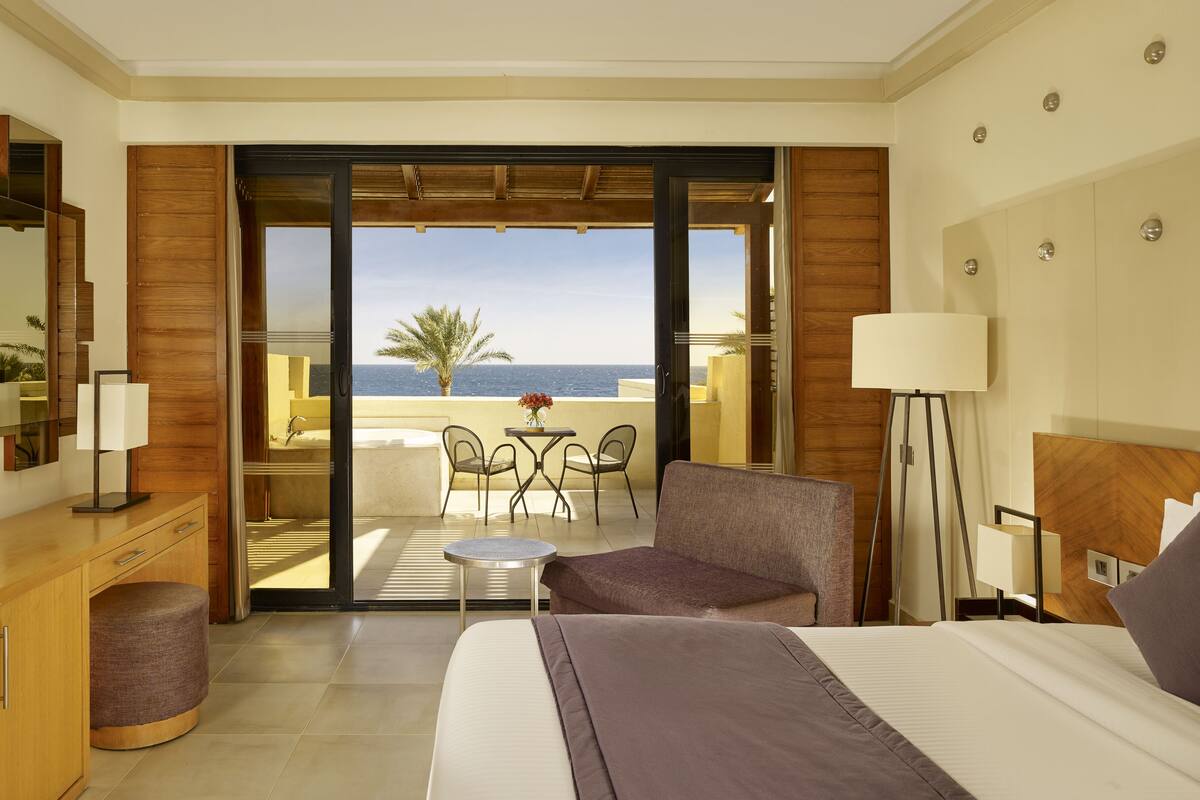 A hotel room with a bed, a desk, a chair, a small table, and a view of the sea.
