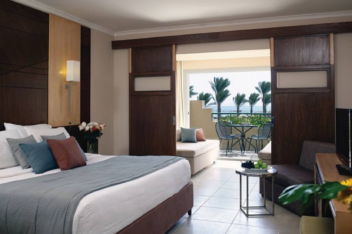 A hotel room with a large bed, a sofa, a small table with fruit, and a balcony with a view of palm trees and the sea.