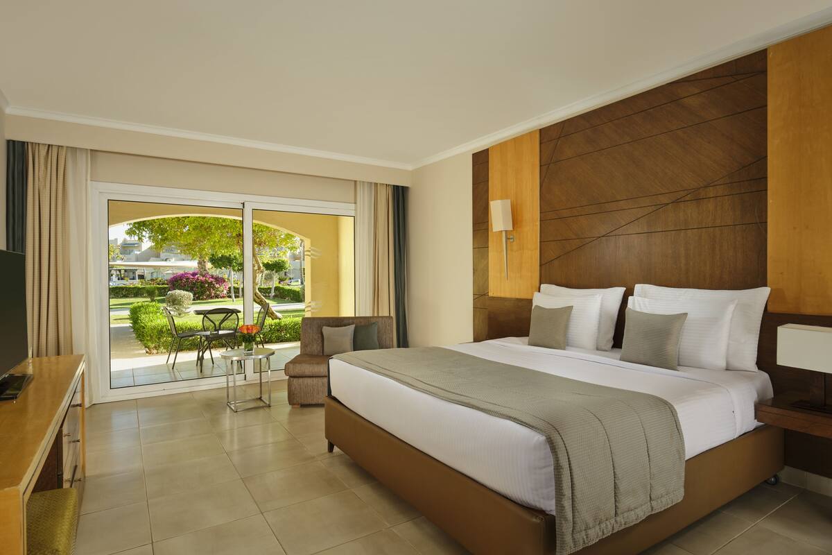 A hotel room with a large bed, a TV, a desk, and a view of a garden through a sliding door.