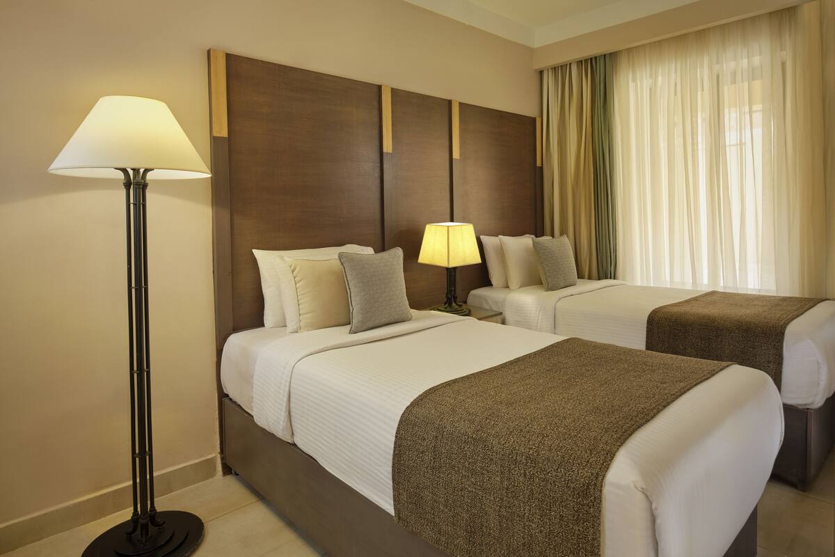 Een hotelkamer met twee bedden, een vloerlamp en een raam met gordijnen.