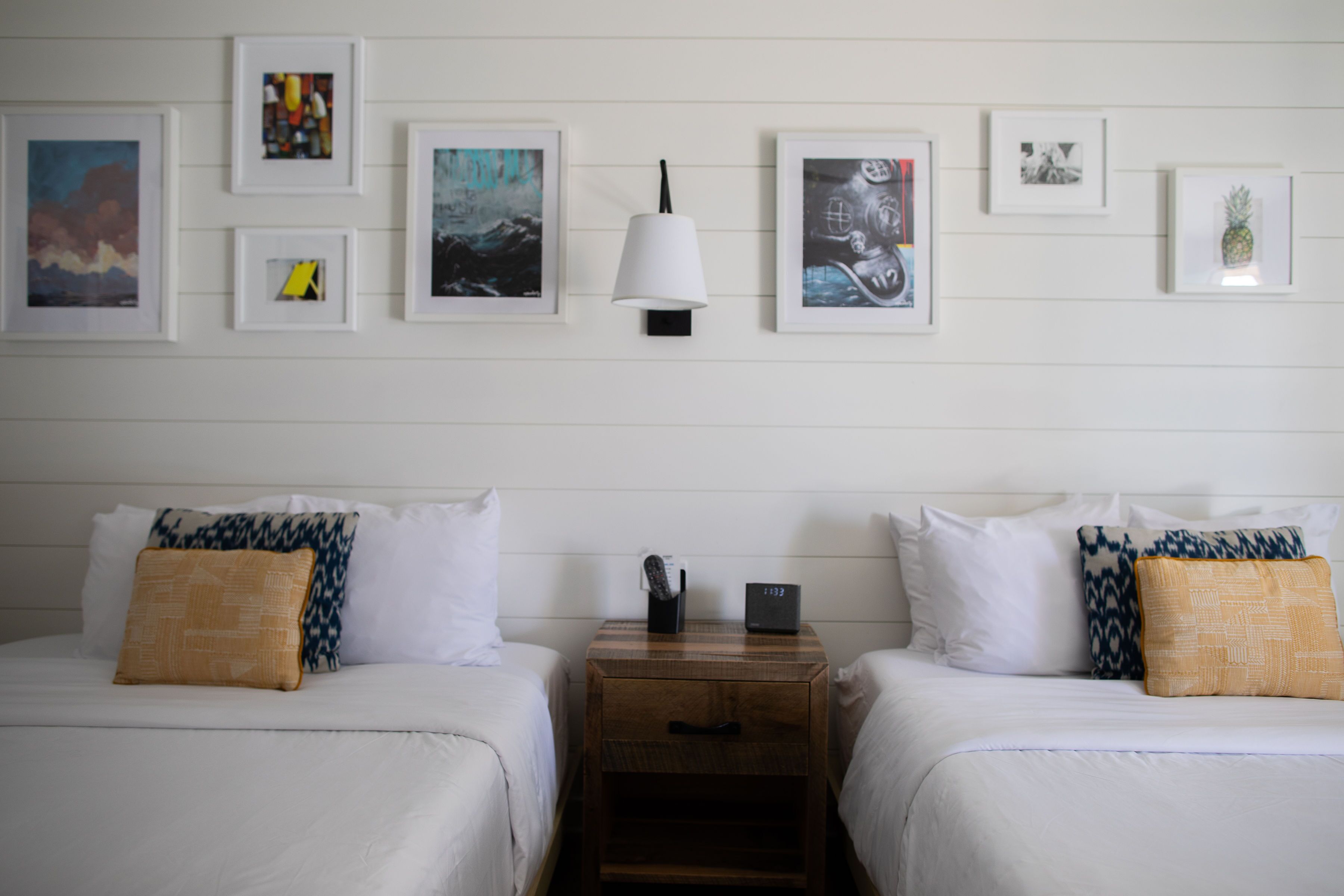 Une chambre d’hôtel avec deux lits, du linge de lit blanc, une table de chevet avec une lampe et des œuvres d’art encadrées accrochées au mur.