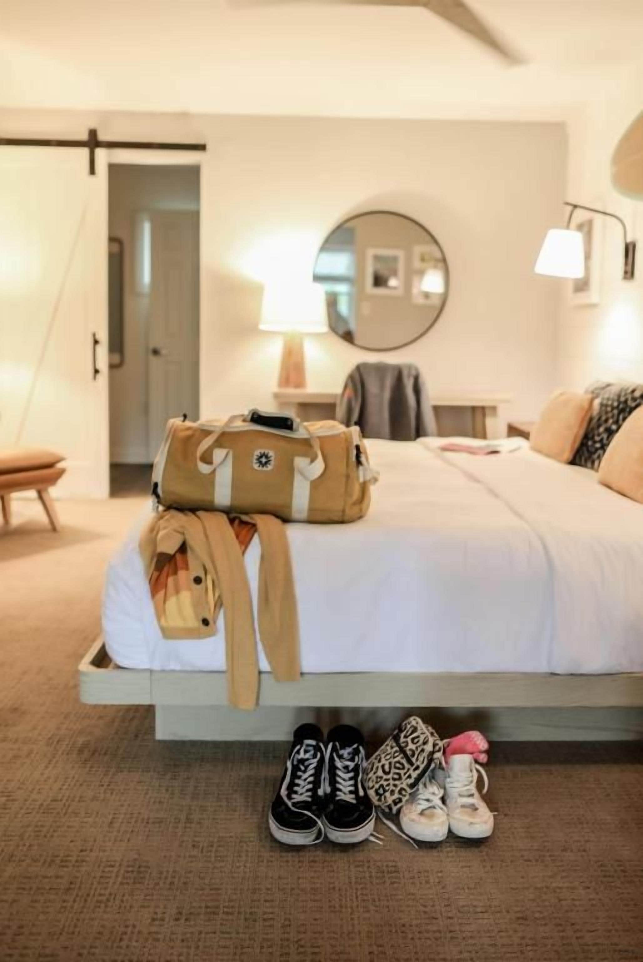 Une chambre d’hôtel avec un lit, une table de chevet, une lampe, un miroir et une valise posée sur le lit.