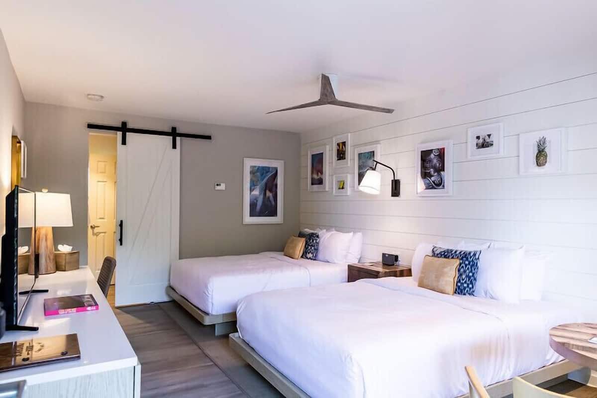 A hotel room with two beds, a desk, a chair, and a ceiling fan.