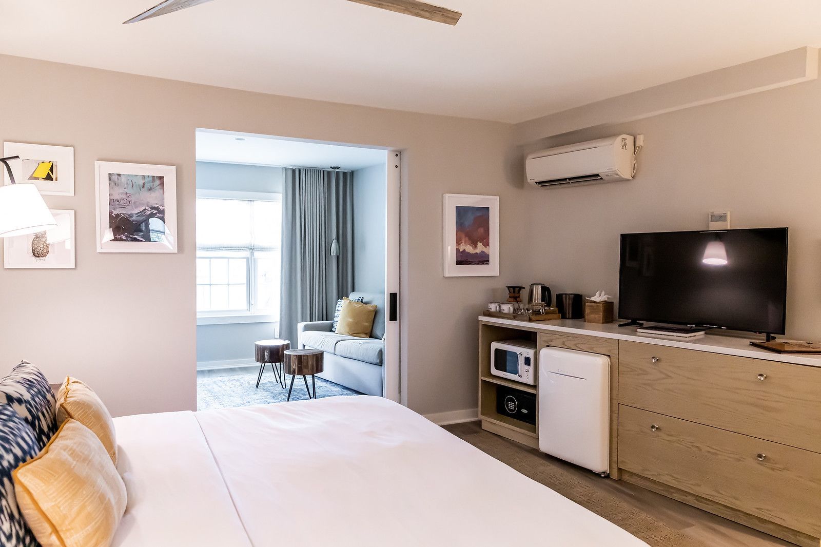 A modern hotel room with a bed, a TV, a kitchenette, and a view of the balcony.