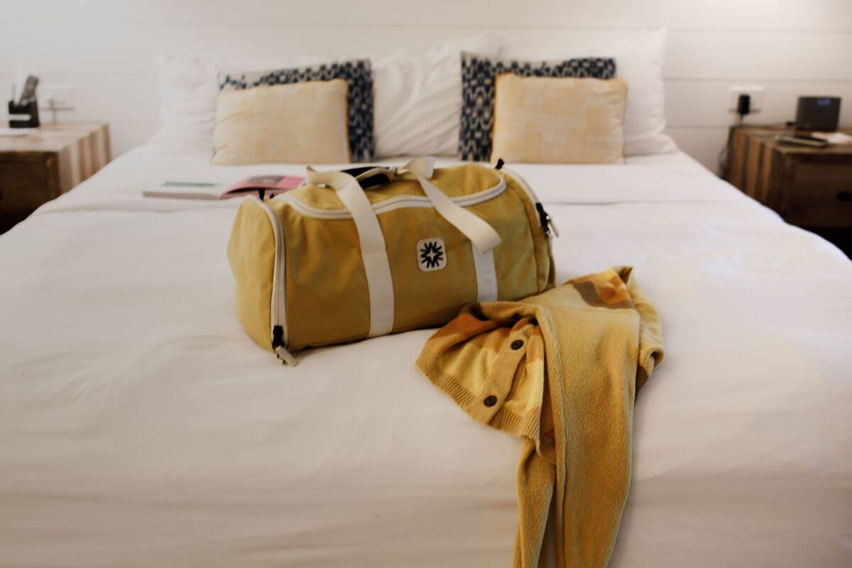 A neatly made bed with a yellow bag and a yellow garment on it. There is a surfboard-shaped sign on the wall above the bed.
