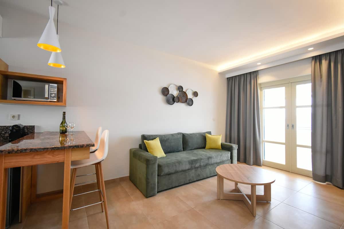 A modern living area with a kitchen, a grey sofa with yellow cushions, a wooden dining table, and a small wooden stool.