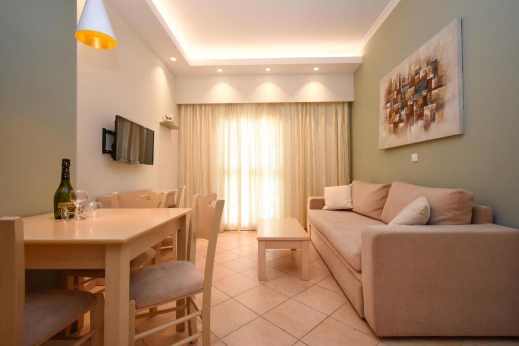 Una sala de estar con un sofá, una mesa de comedor con sillas, un televisor montado en la pared y un ventanal con cortinas.