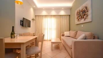 A living room with a sofa, a dining table with chairs, a TV mounted on the wall, and a window with curtains.