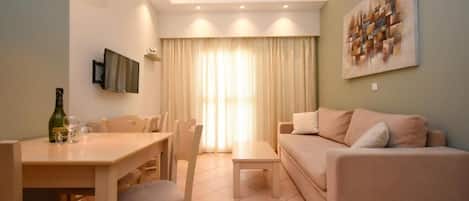 Una sala de estar con un sofá, una mesa de comedor con sillas, un televisor montado en la pared y un ventanal con cortinas.