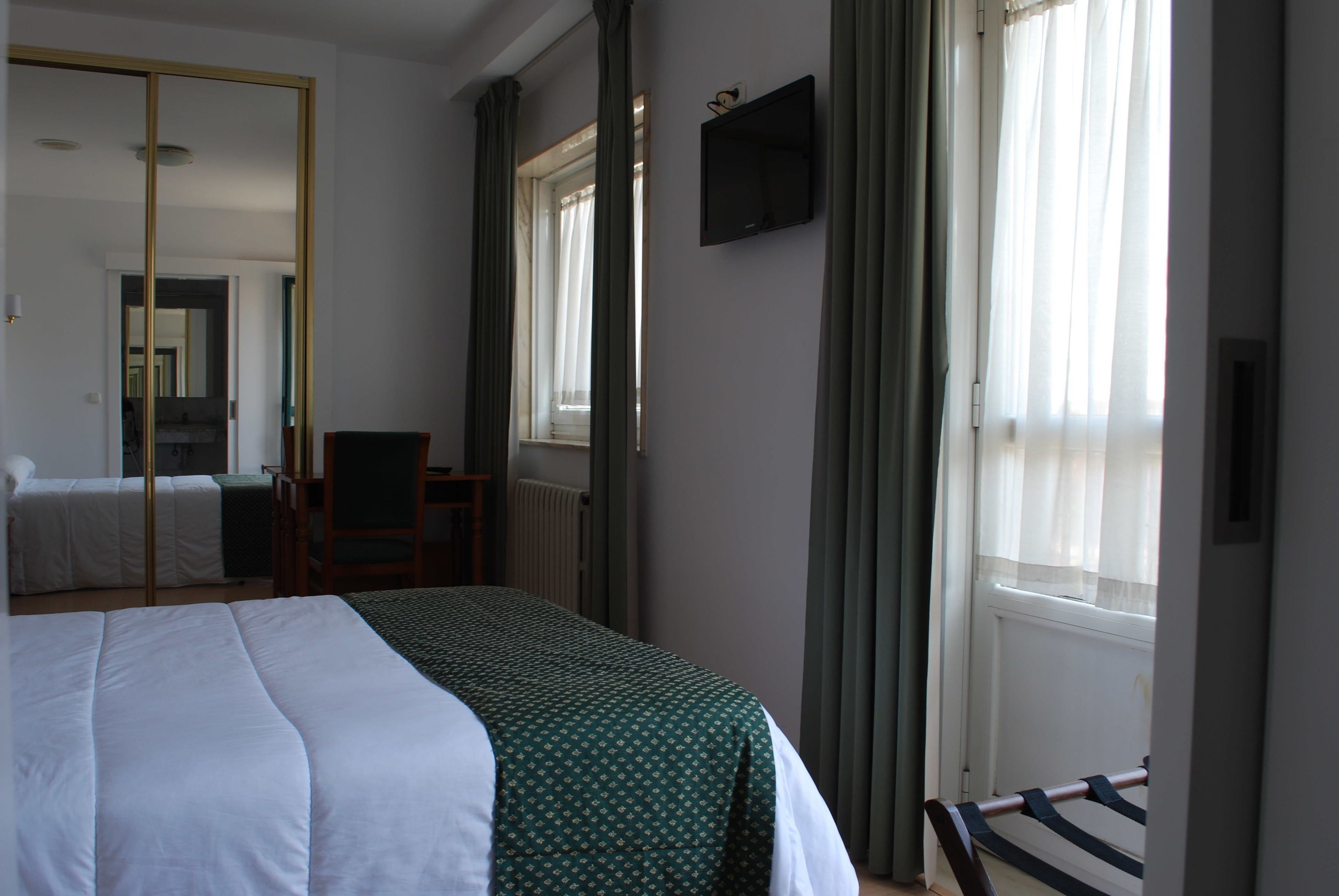 Quarto de hotel com cama, uma cadeira, escrivaninha, televisão e janela com cortinas.