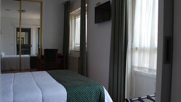Quarto de hotel com cama, uma cadeira, escrivaninha, televisão e janela com cortinas.