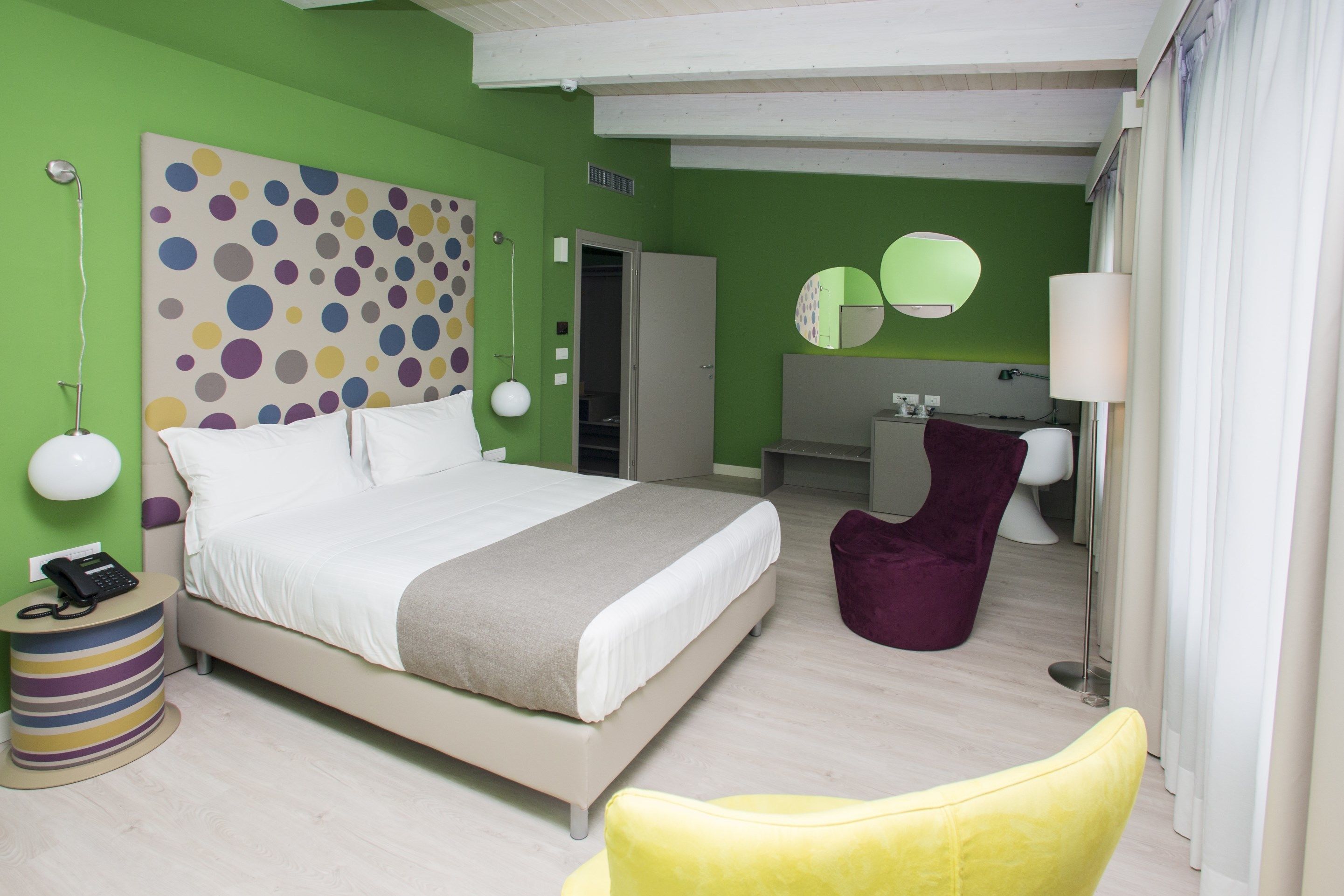 Un dormitorio moderno con una cama grande, un teléfono en la mesita de noche, una obra de arte de colores en la pared, un espejo y una silla morada.