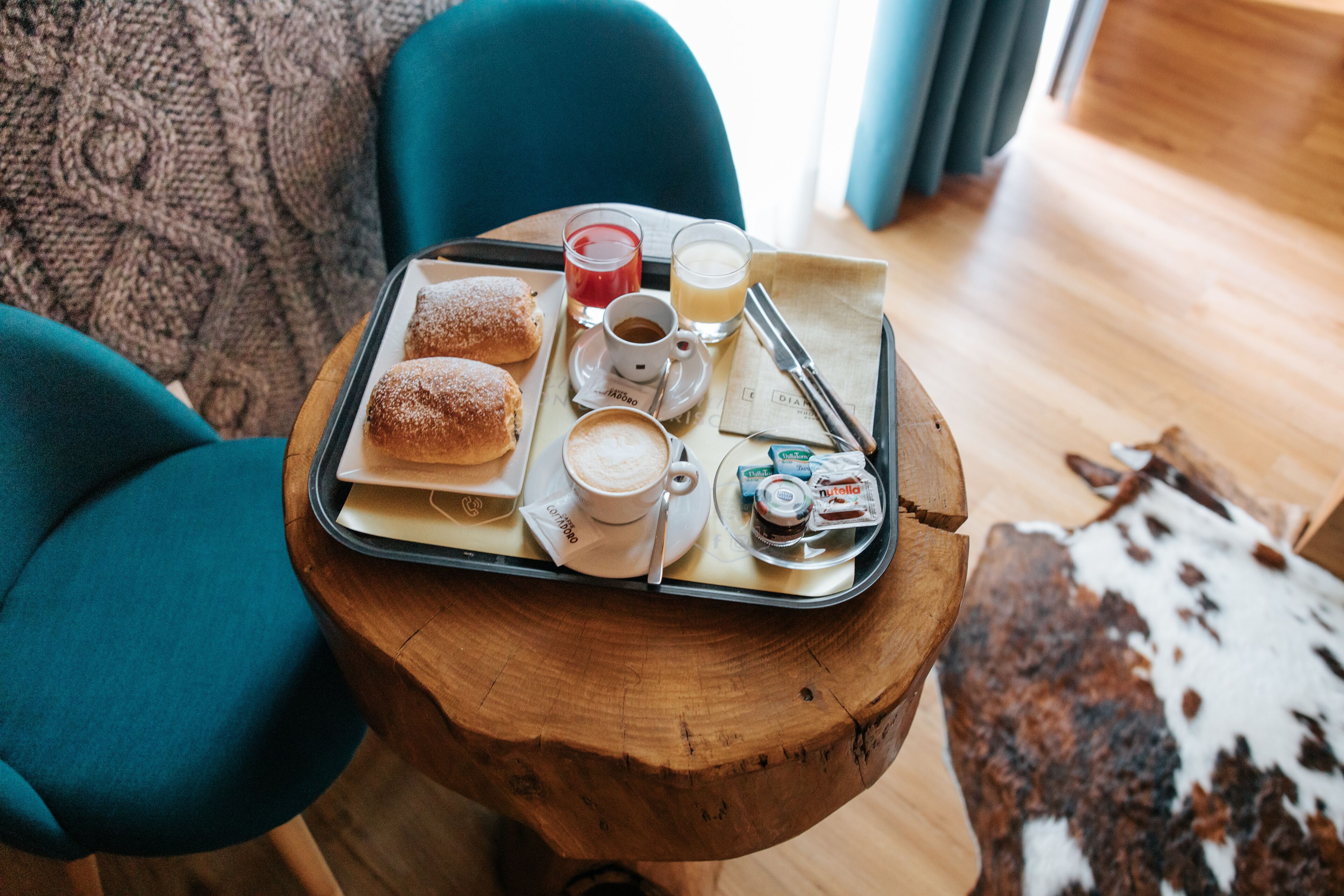 A tray with pastries, coffee, and juice on a wooden table in a cozy room with teal chairs and a cowhide rug.