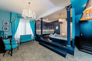 Quarto de hotel moderno com uma cama grande, um espaço de estar com sofá e poltronas, um candelabro e vista para a cidade.