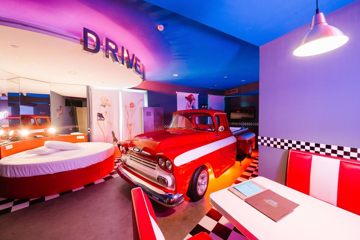 A vintage-themed room with a classic red car, a bed, and a diner-style table and booth.
