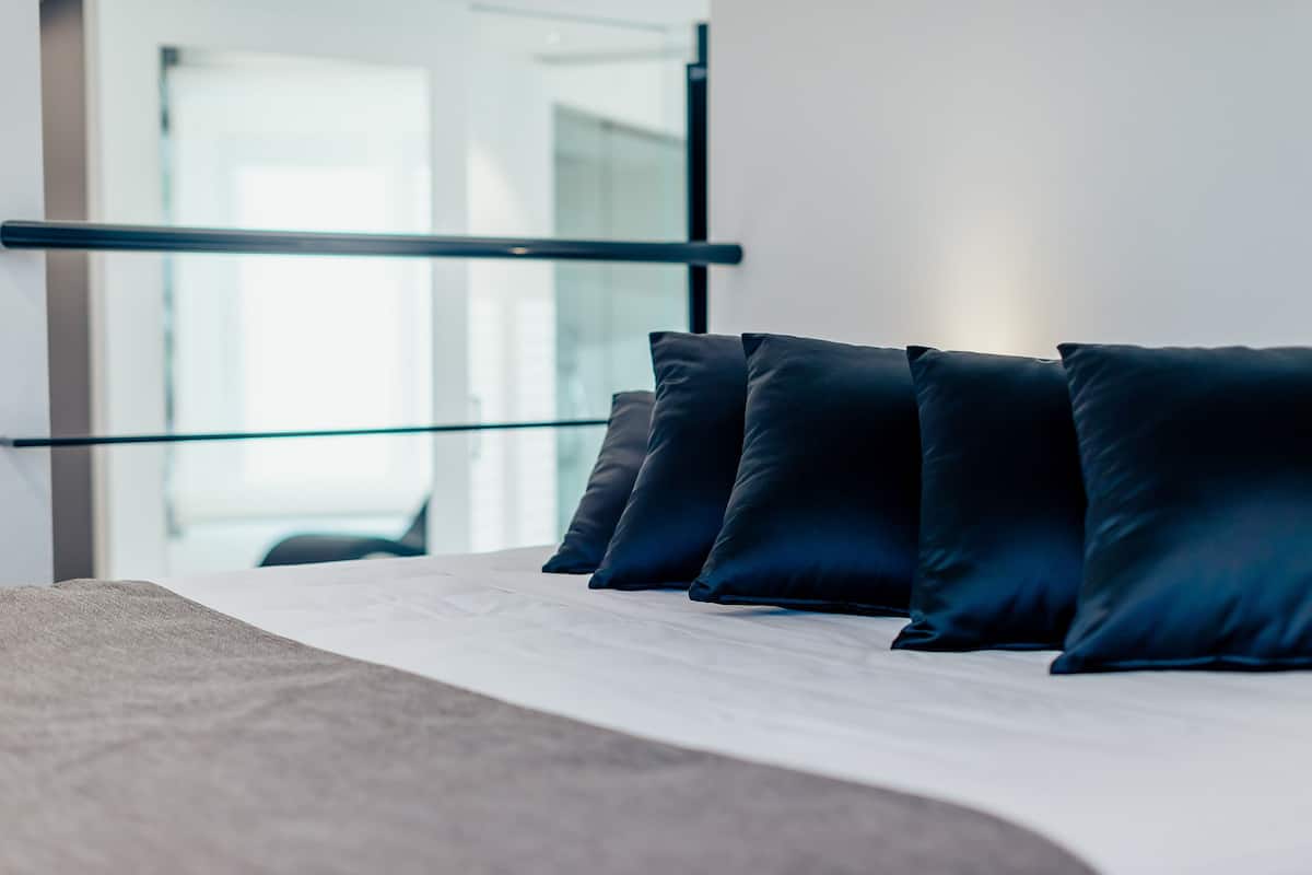 A bed with dark pillows and a white mattress, a glass railing, and a modern interior.