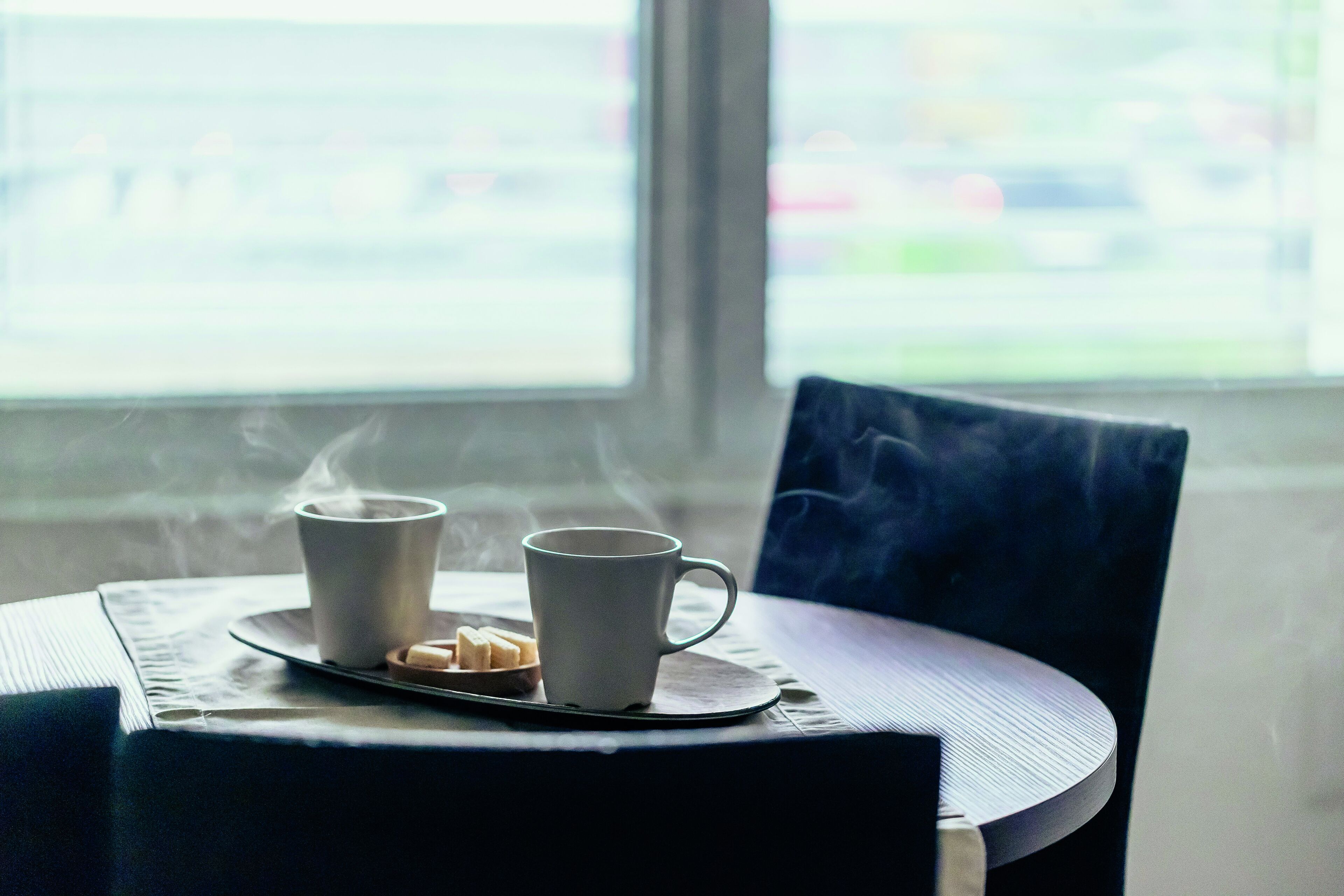 Deux tasses blanches, posées sur un sous-verre, d’où s’échappe de la vapeur, ce qui laisse supposer qu’elles contiennent des boissons chaudes. Elles sont placées sur une table ronde, à côté d’une chaise sombre.