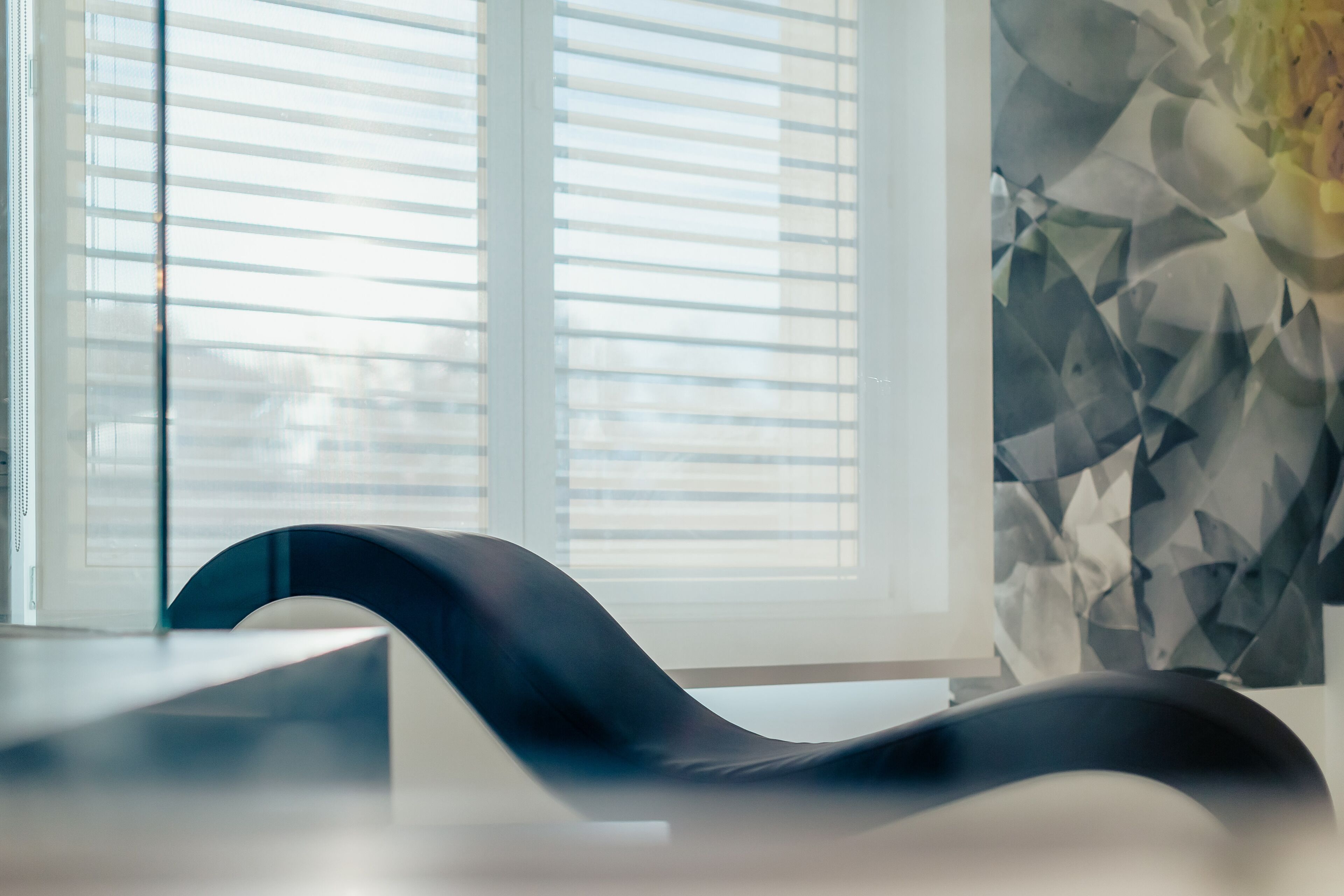 Ein modernes Zimmer mit einem schwarz gebogenen Sessel, einem Fenster mit Jalousien und einer Wand mit geometrischem Muster.