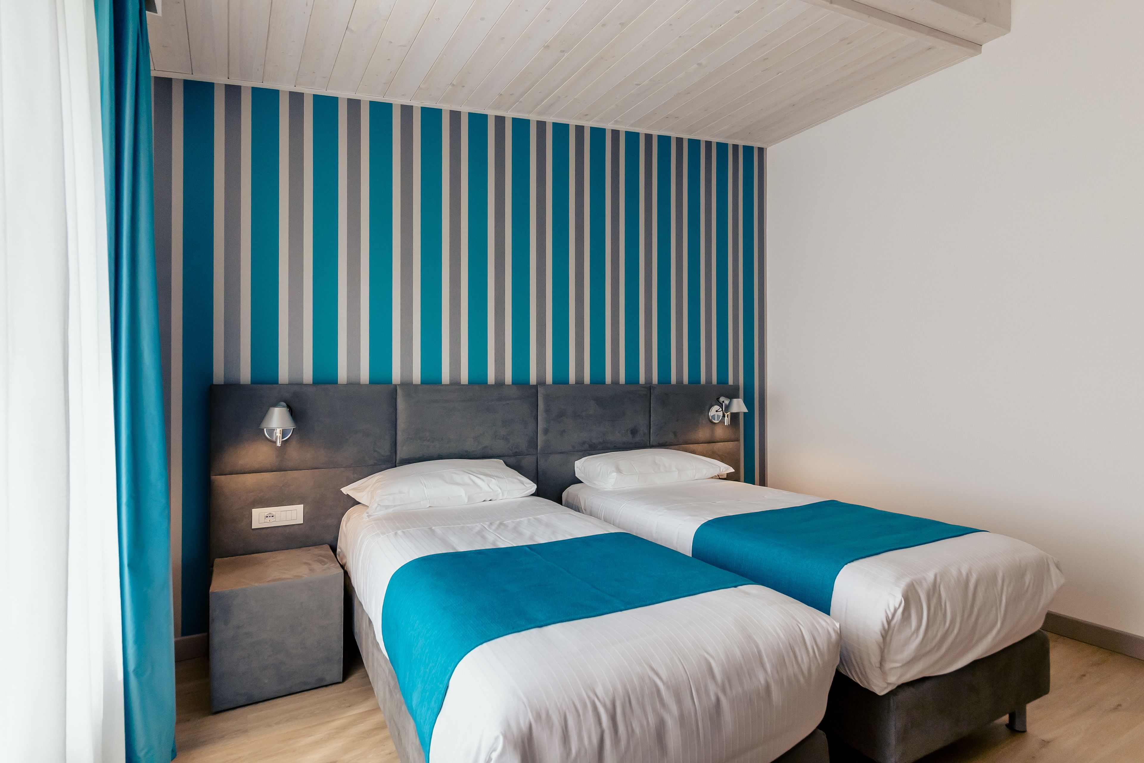 A modern hotel room with a striped wall, two beds, a nightstand, and a ceiling with wooden beams.