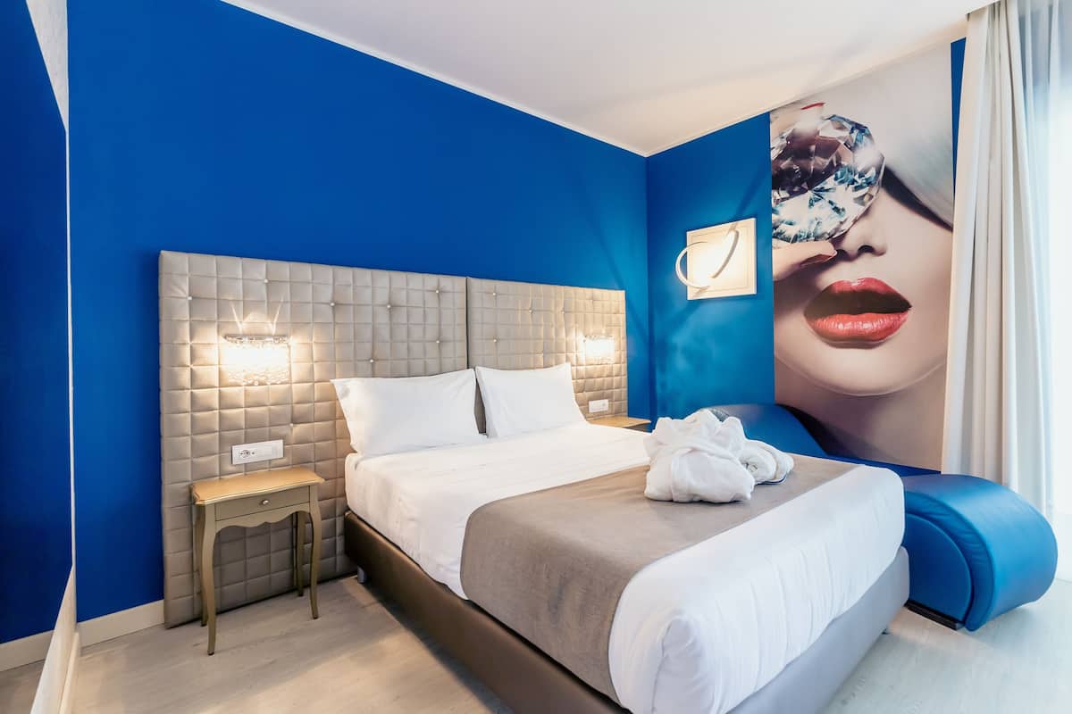 A modern hotel room with a blue accent wall, a tufted headboard, a bedside table with a lamp, and a large poster on the wall.