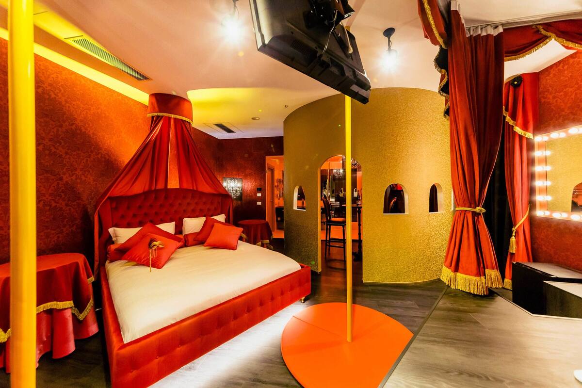 A room with a large bed, a red canopy, a mirror with lights, and a small table with a red tablecloth.