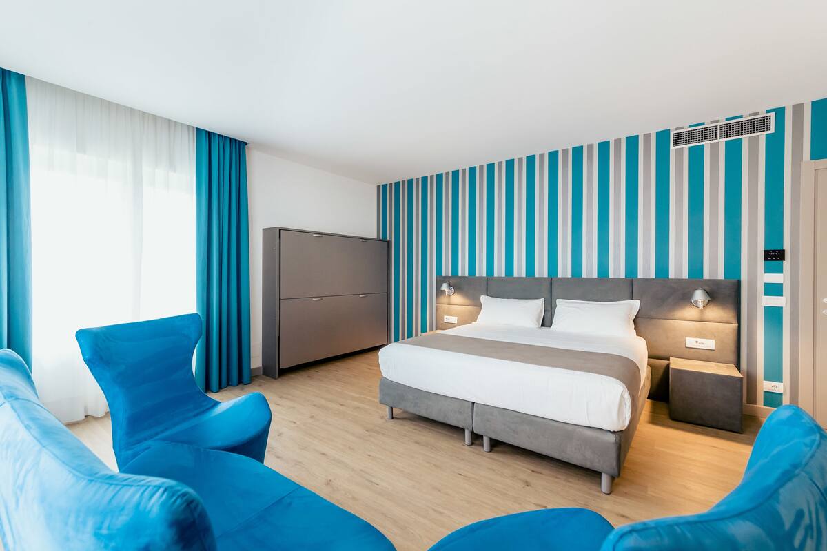 A modern hotel room with a bed, two bedside tables, a wardrobe, and a seating area with blue chairs.