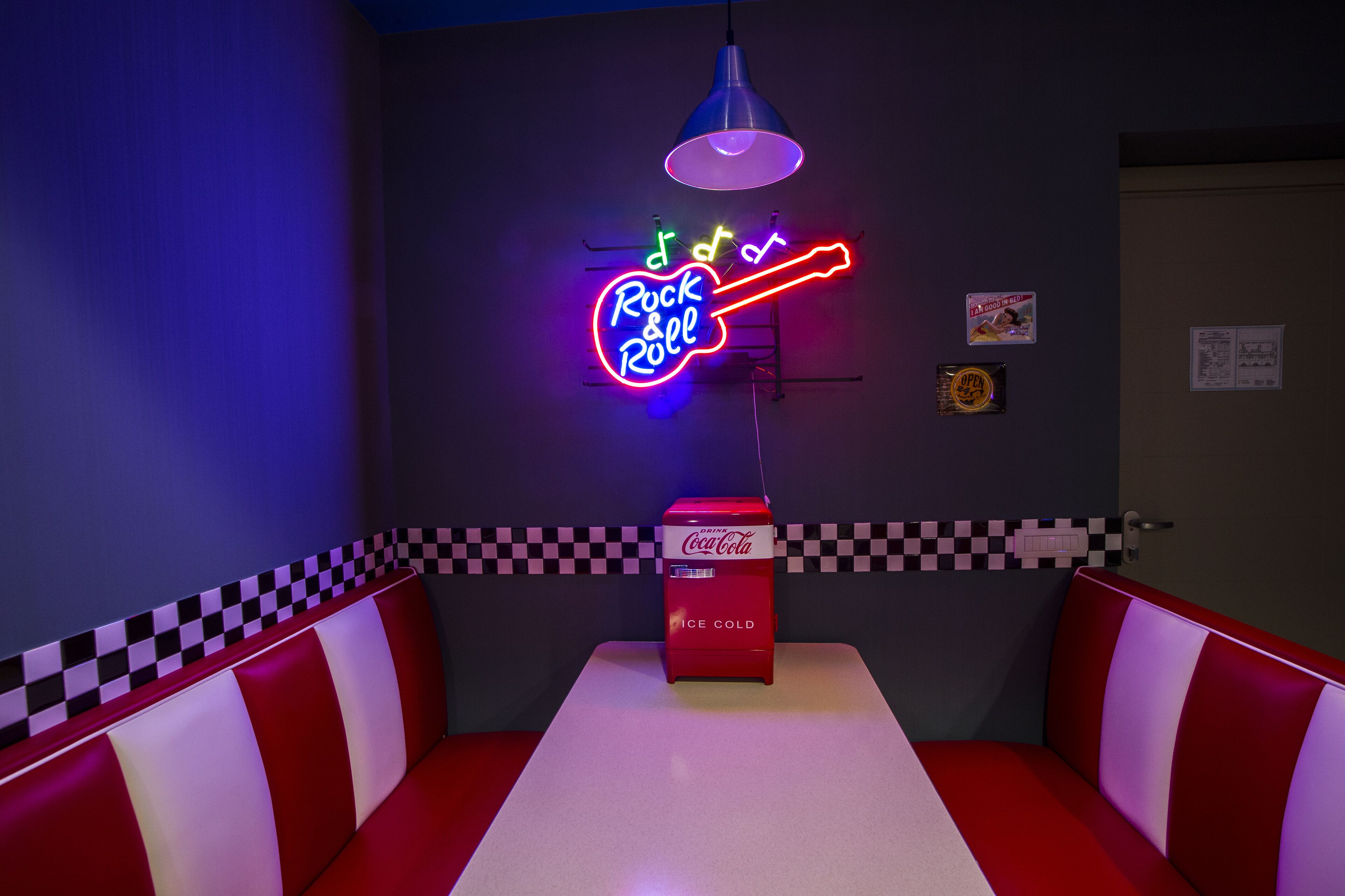 A neon sign reading "Rock & Roll" above a Coca-Cola vending machine in a diner setting with checkered wall and red and white striped booths.