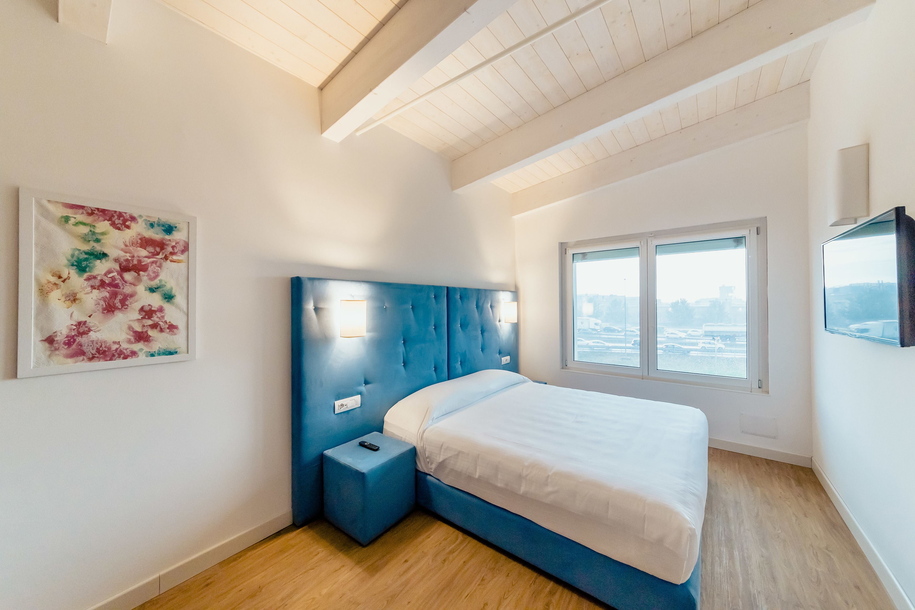 A bedroom with a blue headboard, a bedside table, a bed with white bedding, a window with a view, and a framed artwork on the wall.