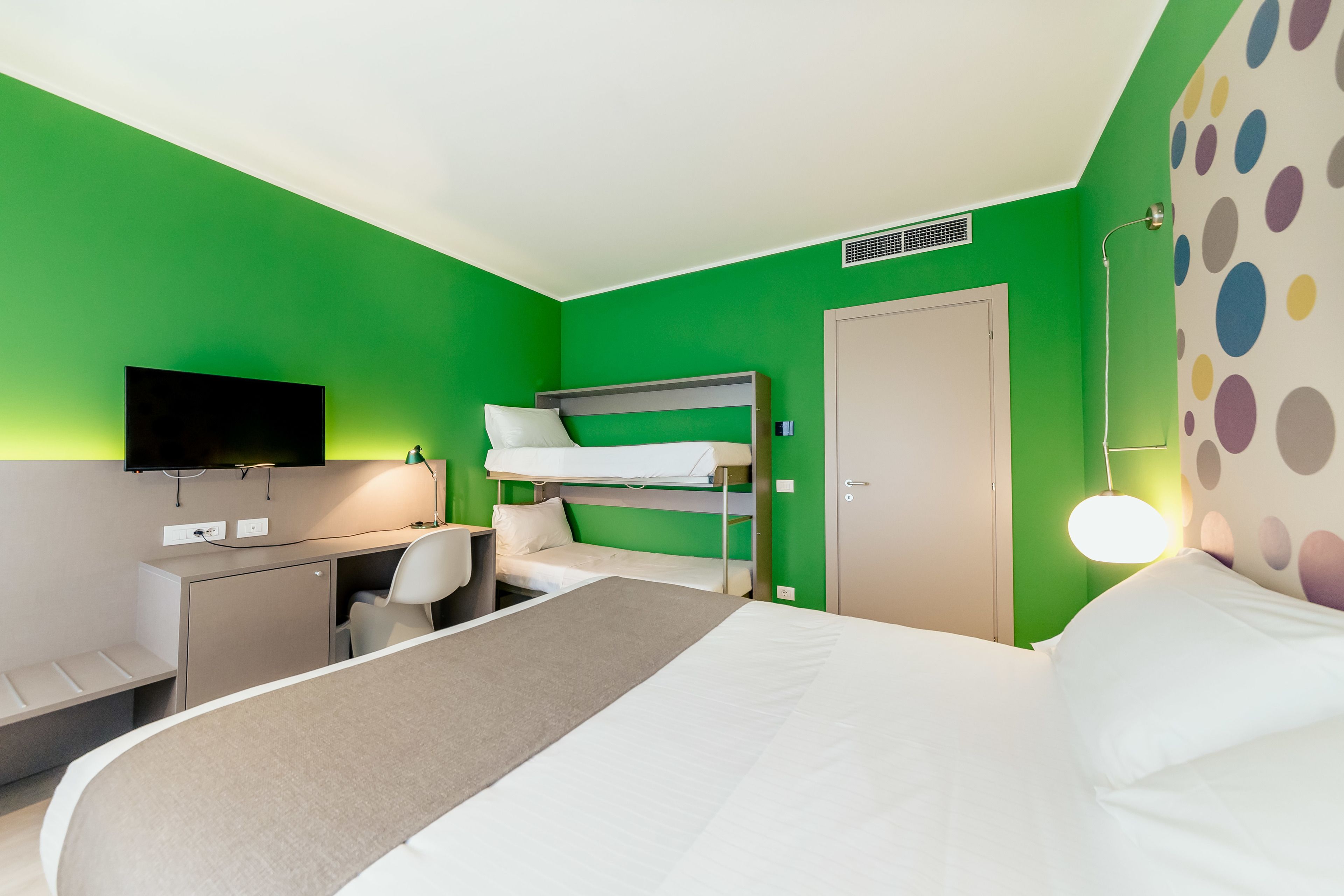 A hotel room with a bunk bed, a desk with a chair, a television, and a bed with a pillow.