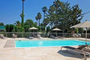 A swimming pool with lounge chairs and umbrellas, surrounded by palm trees and a fence. - Hotel Poretta (Lucciana)
